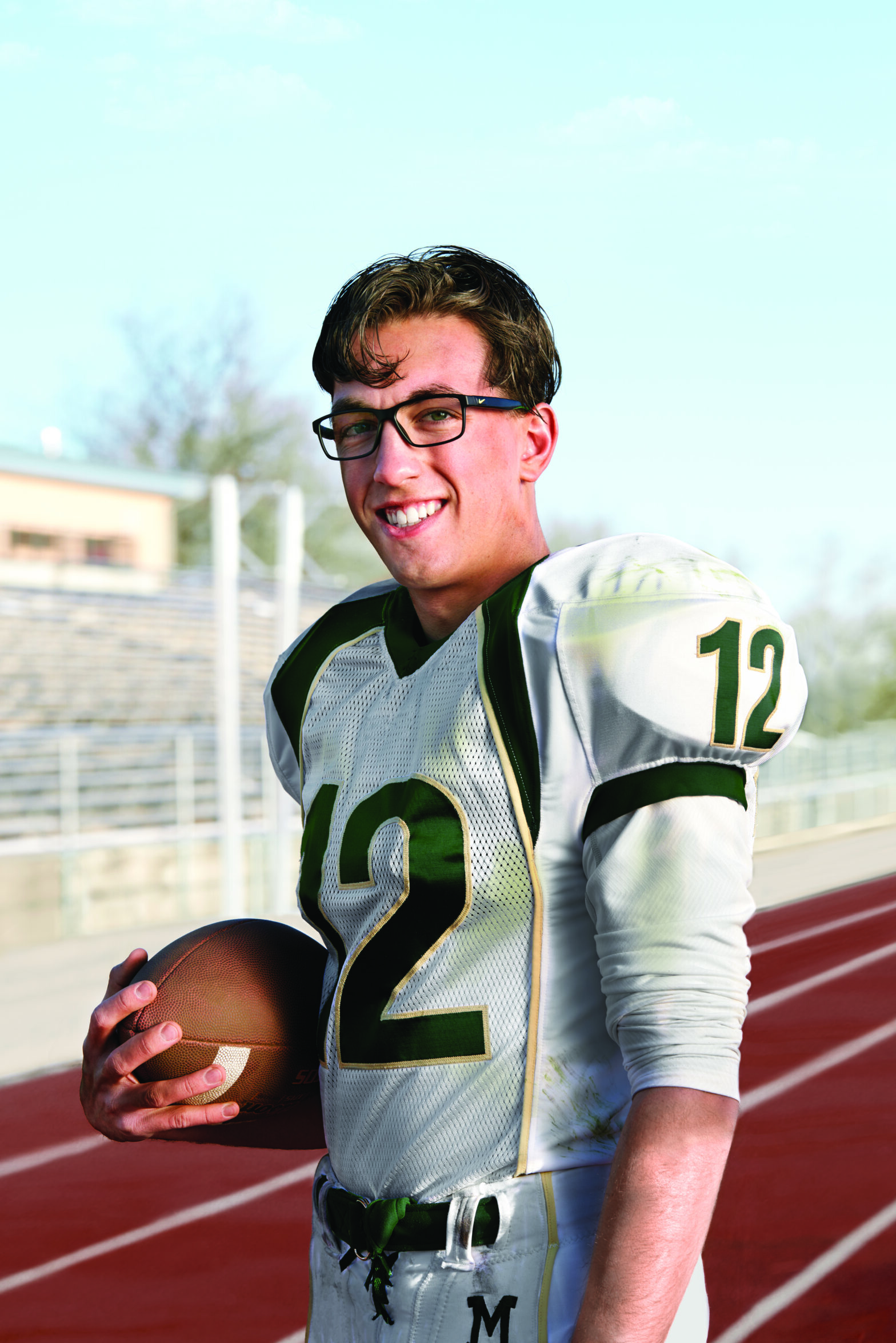 An athlete holds a football and poses in his uniform and black-framed glasses.