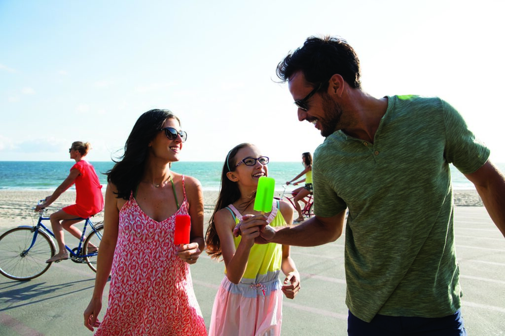 A family wearing glasses and sunglasses enjoy popsicles near a sunny beach.