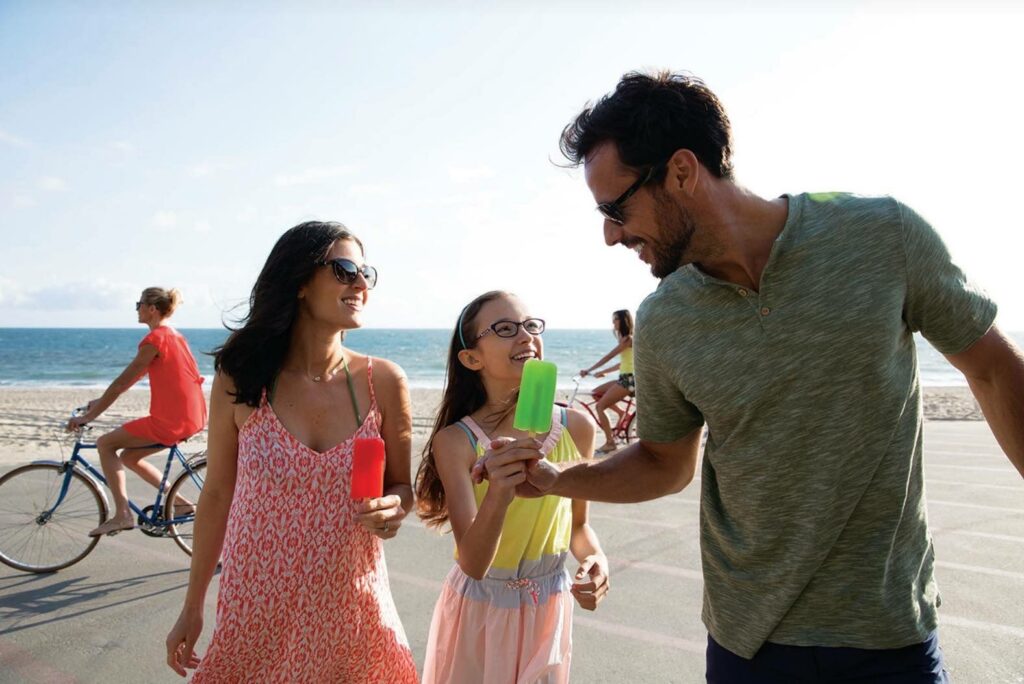A smiling man in sunglasses hands a green popsicle to his daughter as his wife stands to the side, smiling and holding a red popsicle. A sunny beach and two bicyclists can be seen in the background.