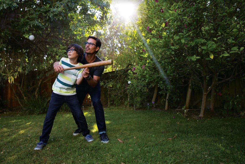 A father helps a son, both wearing glasses, swing a bat at a baseball in a sunny, wooded area.