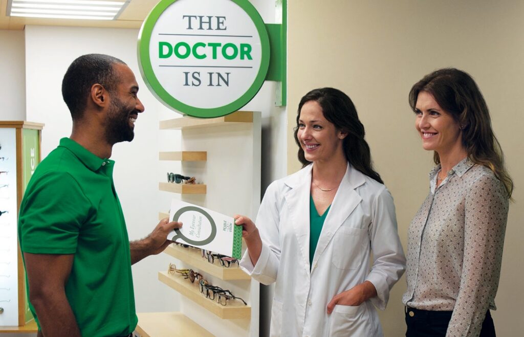 A smiling woman in a white lab coat hands a brochure to a smiling man on her right while a woman on her left looks on, smiling.