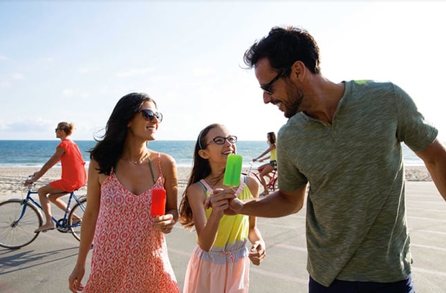 A husband, wife and daughter, all in glasses, enjoy popsicles on the beach.