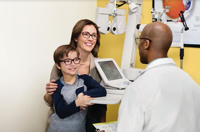 A mother and son with glasses smile in a vision care franchise exam room with an optometrist.