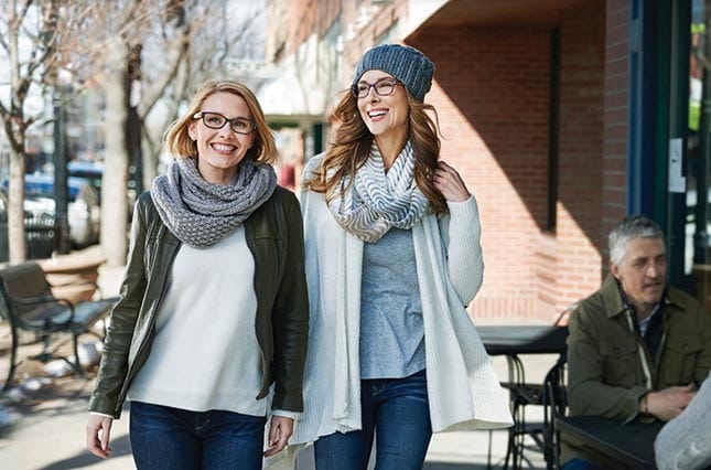 Two women wearing stylish glasses walk past a cafe.