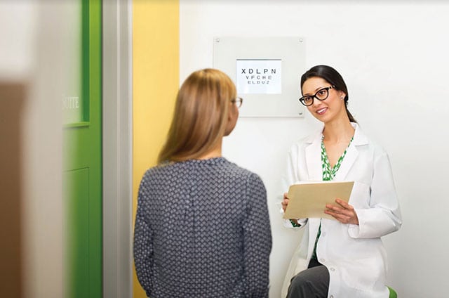 A female optometrist works with a female patient who is reading an eye chart on the wall.