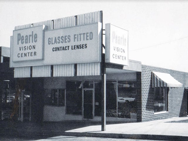 A black-and-white photo of a vintage Pearle Vision corner store. The sign in front says “Pearle Vision Center. Glasses Fitted. Contact Lenses.”