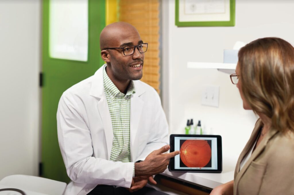 An optometrist points at an image of an eye on a tablet while facing his patient, who is to the right of the frame and slightly out of focus.