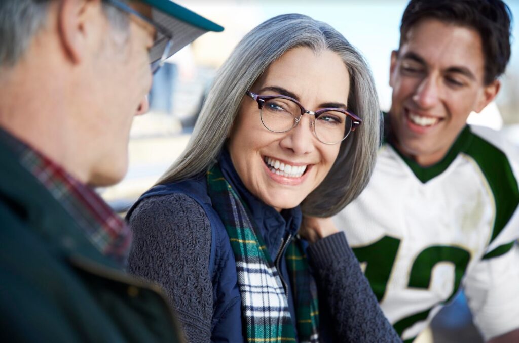 A smiling older woman is wearing glasses as her husband sits to her right and a grandson in a football uniform sits to her left, both slightly out-of-focus.
