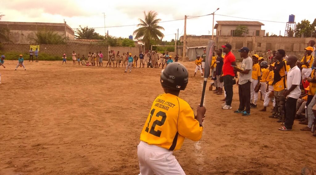 A young batter in a black helmet and yellow baseball uniform stands with his bat, ready for an incoming pitch, on a baseball field in Benin as onlookers and teammates watch.