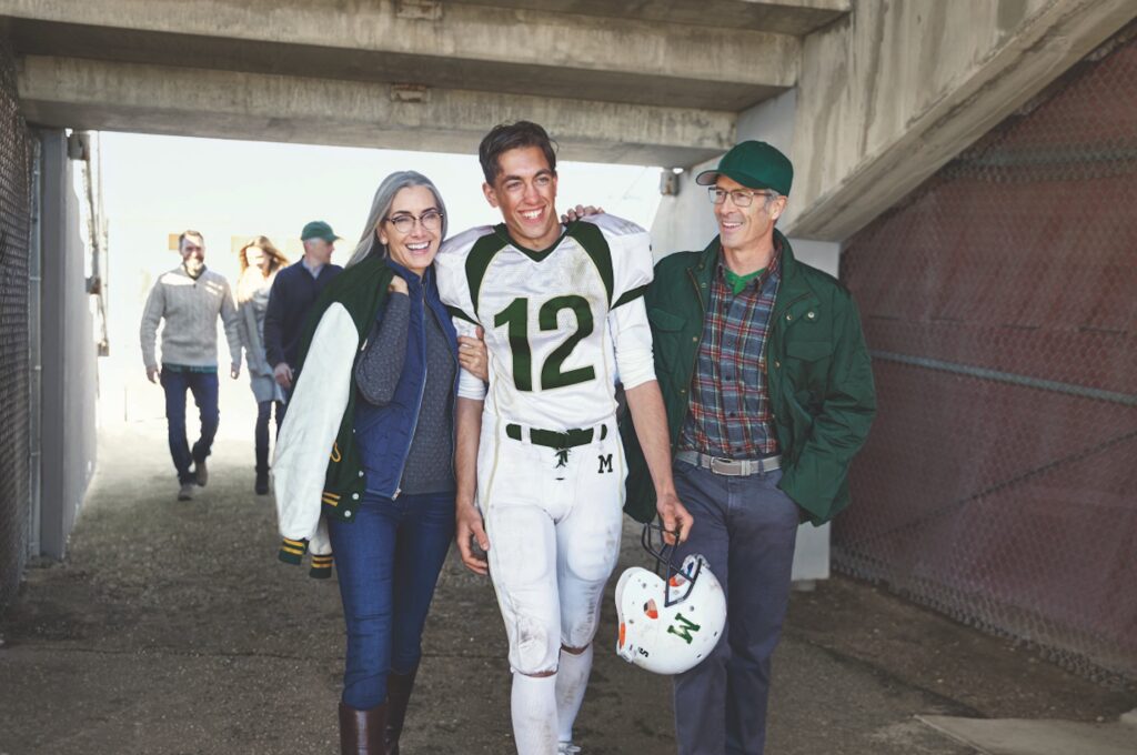 A young football player wearing the number 12 on his jersey in green is flanked by an older woman on his left and an older man on his right, both of whom are smiling and wearing glasses.