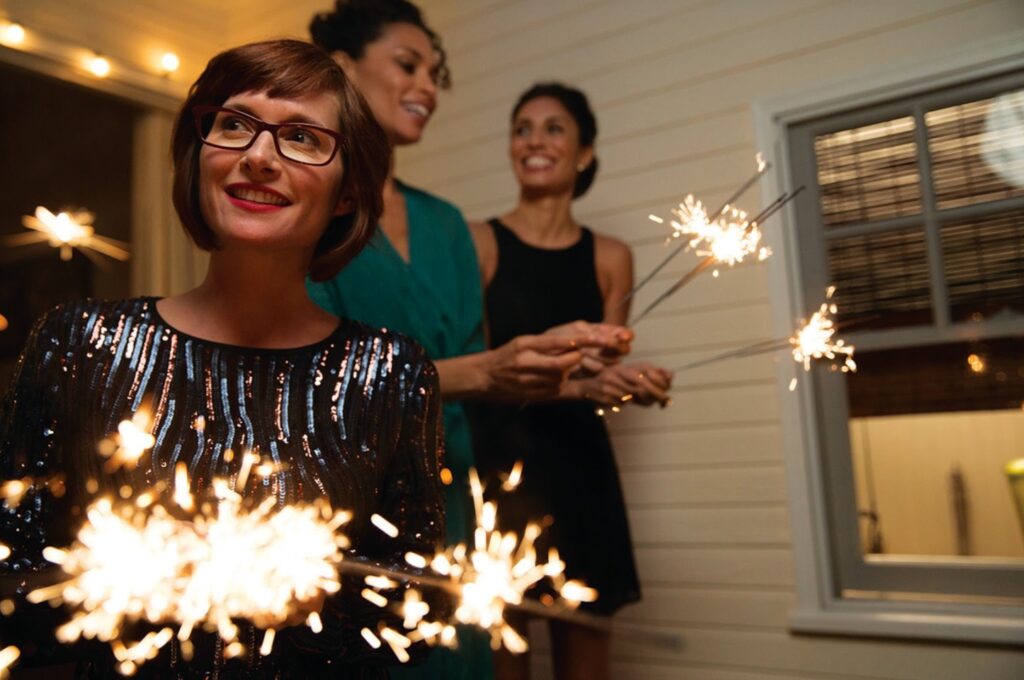 A woman wearing glasses and two out-of-focus women in the background hold sparklers at night on a house’s porch.
