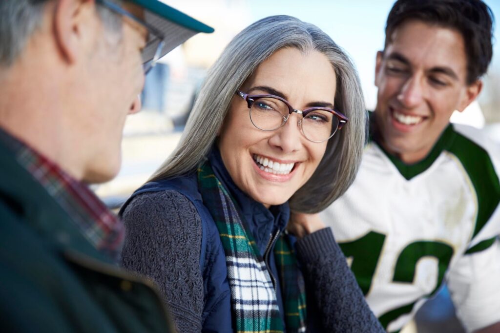 A silver-haired woman in glasses smiles toward the camera; her husband is visible in the foreground and her son in the background in a football uniform.