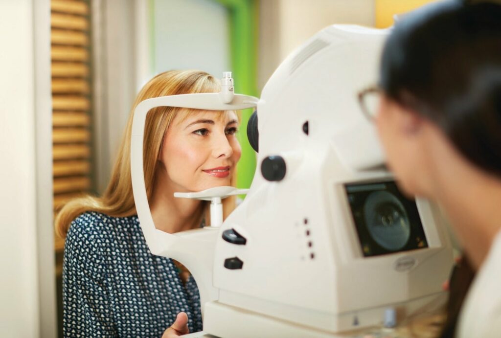 A Pearle Vision patient rests her chin on an eye exam machine; the machine’s operator is visible but out of focus in the foreground.