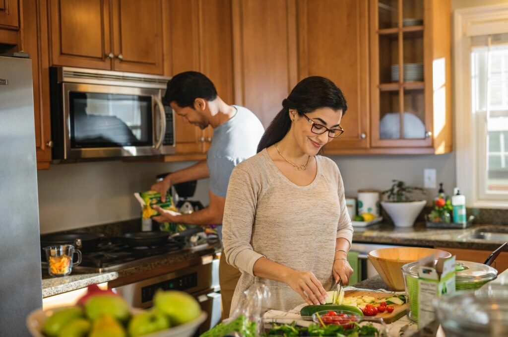 A smiling woman in glasses chops vegetables in her kitchen; visible but slightly out-of-focus in the background, her husband is pouring food into a skillet.