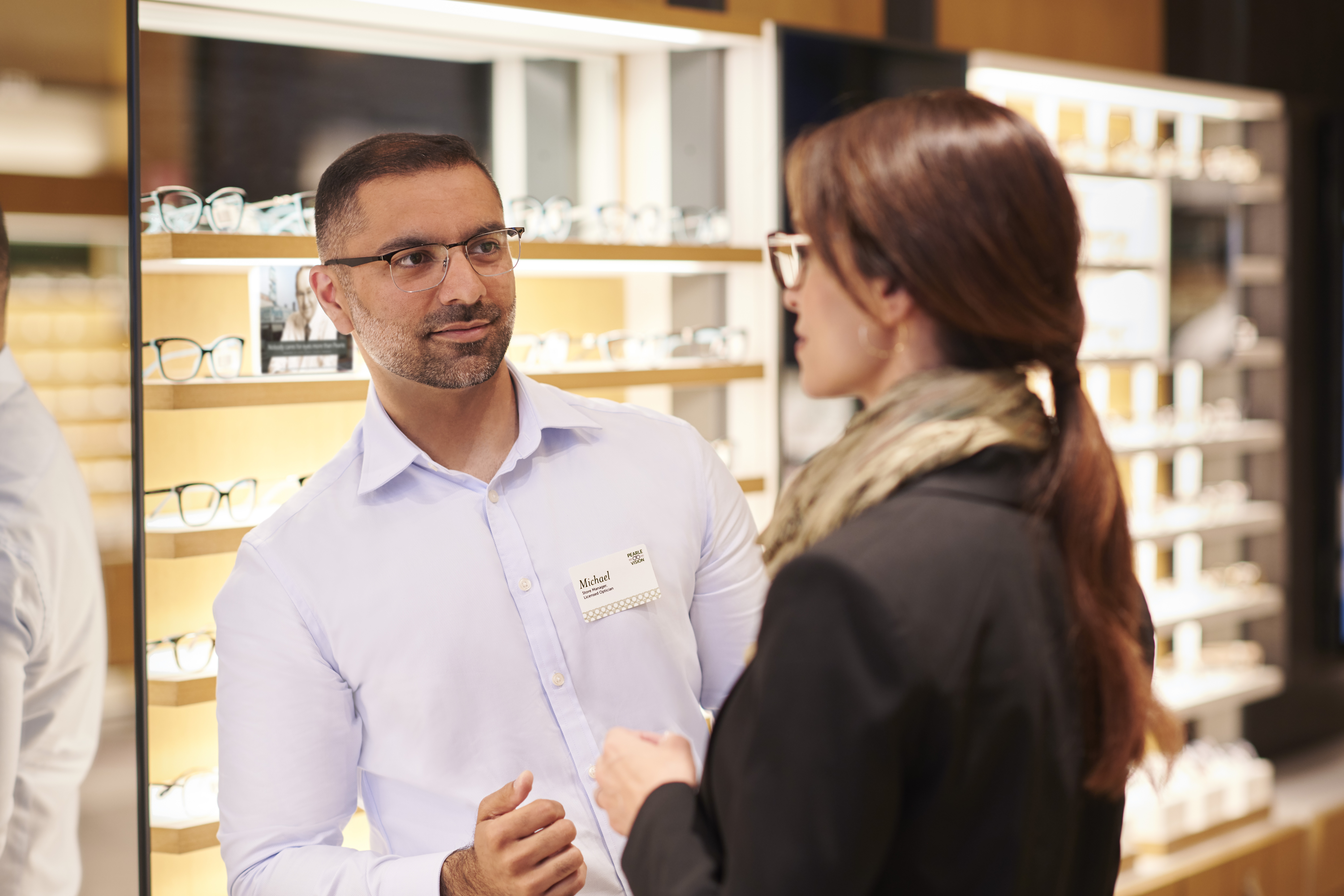 A customer smiles while trying on a pair of glasses in a Pearle Vision EyeCare Center. A store associate is also pictured out-of-focus in the foreground.