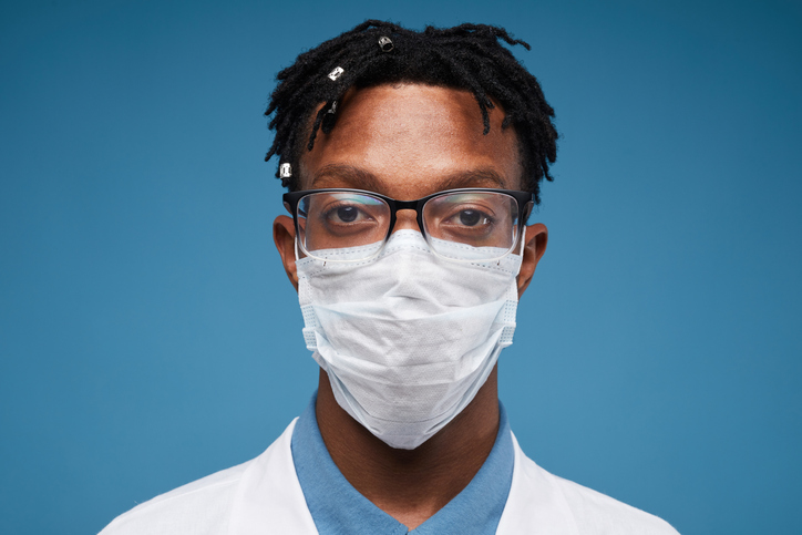 Head and shoulders portrait of optometrist wearing protective mask and looking at camera while posing against blue background